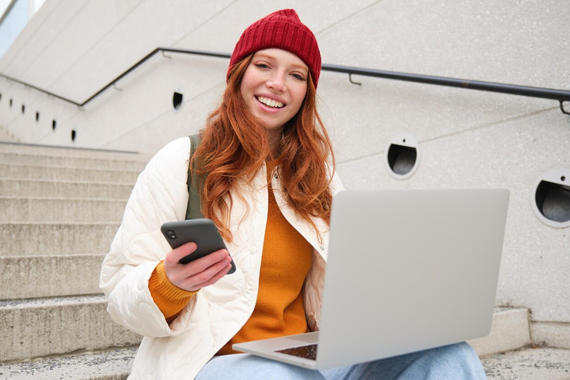 Junge, freundliche, weibliche Person sitzt draußen auf einer grauen Treppe. Sie hat einen Laptop auf ihren Beinen und in der rechten Hand ein Handy. Sie hat rote Haare und trägt einen orangefarben Pulver mit einer weißen Jacke, sowie eine blaue Jeans und eine rote Mütze.